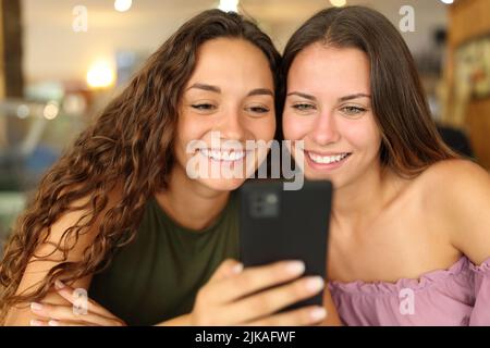 Zwei fröhliche Frauen, die lächelnd in einer Bar ihr Smartphone checken Stockfoto
