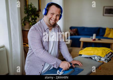Unabhängiger junger Mann mit Down-Syndrom, der zu Hause Musik hört und Kleidung bügelt. Stockfoto