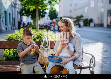 Junge Mutter mit kleinen Kindern, die sich auf der Bank in der Stadt ausruhen und einen Obstsnack essen. Stockfoto