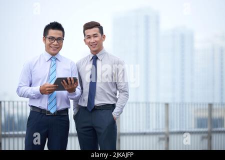 Zwei glückliche vietnamesische Buisnessmen stehen draußen, große Stadt ist im Hintergrund Stockfoto