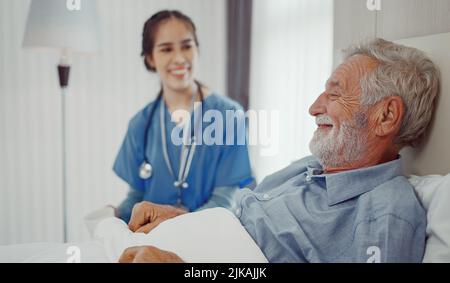 Krankenschwester, die im Zimmer auf der Krankenstation einen älteren Mann ins Bett bringt. Stockfoto