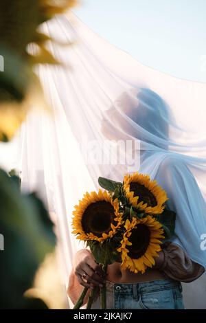 Unbekannte Frau mit Sonnenblumenstrauß mit weißem Stoff im Freien bedeckt. Natürliche Schönheit. Ländlicher Sommerurlaub, Reinheit und Naturkonzept. Speicherplatz kopieren Stockfoto