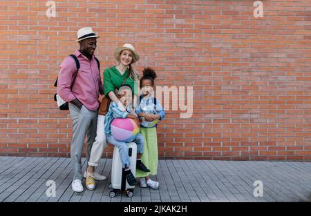 Multirassische Familie, die zusammen mit kleinen Kindern reist. Posiert vor einer Ziegelwand. Stockfoto