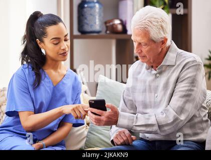 Helfen Sie mir dabei? Eine junge Frau, die ihrem älteren Patienten hilft, benutzt ein Smartphone. Stockfoto