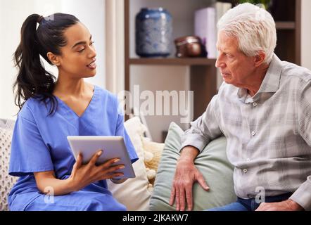 Eine junge Frau, die ihrem älteren Patienten hilft, benutzt ein digitales Tablet. Stockfoto