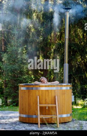 Junge Frau, die in der Holzbadewanne mit Kamin Holz verbrennt und Wasser im Hinterhof in den Bergen erwärmt. Stockfoto