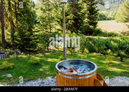 Junge Frau, die in der Holzbadewanne mit Kamin Holz verbrennt und Wasser im Hinterhof in den Bergen erwärmt. Stockfoto