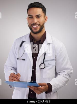 Wie fühlen Sie sich heute. Ein hübscher junger Arzt, der allein im Studio steht und ein Klemmbrett hält. Stockfoto