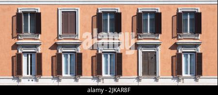 An einem sonnigen Tag in Rom, Italien, wurden die Fenster geschlossen, einige geschlossen, in einem Mehrfamilienhaus mit einer orangefarbenen verputzten Wand. Breitbild Stockfoto
