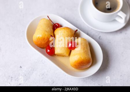 Neapolitanischer Rum baba (oder baba au Rum) auf einem weißen Teller mit einer Cocktailkirsche und einer Tasse Kaffee. Kleine, in Rumsirup getränkte Hefekuchen auf einem grauen Rücken Stockfoto