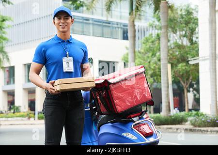 Fröhlicher, hübscher junger Lieferer, der Kisten mit heißer Pizza hält Stockfoto