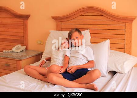Zwei Kinder liegen im Urlaub in einem Hotelzimmer auf einem Bett mit weißen Laken. Kinder spielen zu Hause auf dem Bett mit weißen Laken. Junge und Mädchen, bevor sie zu Hause ins Bett gehen Stockfoto