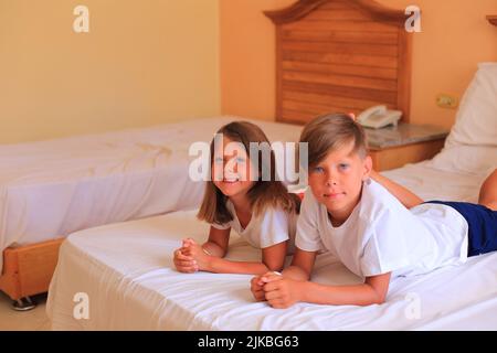 Zwei Kinder liegen im Urlaub in einem Hotelzimmer auf einem Bett mit weißen Laken. Kinder spielen zu Hause auf dem Bett mit weißen Laken. Junge und Mädchen, bevor sie zu Hause ins Bett gehen Stockfoto