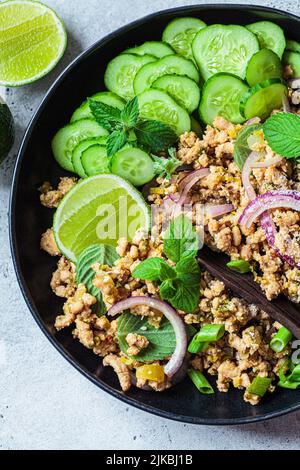 Thailändischer Hähnchensalat, Larb Gai mit Zwiebel, Gurke und Minze auf einem schwarzen Teller, Nahaufnahme. Asiatisches Speisekonzept. Stockfoto