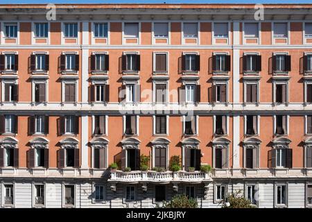 Apartmentgebäude mit Balkonen und verschlossenen Fenstern, einige geschlossen, an einem sonnigen Tag in Rom, Italien, in einer orangefarbenen verputzten Wand. An der Spitze einer Borde Stockfoto
