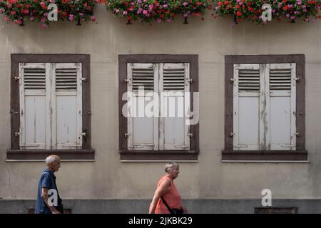 Mann und Frau gehen an einer alten Fassade mit Fensterläden und Blumenkästen in der Grand-Rue in der alten französischen Stadt Ribeauvillé im Elsass vorbei Stockfoto