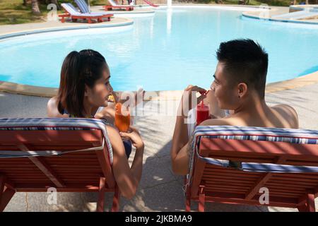 Glückliches junges vietnamesisches Paar, das am Pool liegt und Smoothies trinkt Stockfoto