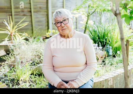 Porträt einer lächelnden älteren, reifen Großmutter im Ruhestand in einer Brille. Glückliche ältere Frau, die lächelt und in ihrem grünen Garten sitzt und den Ruhestand genießt Stockfoto