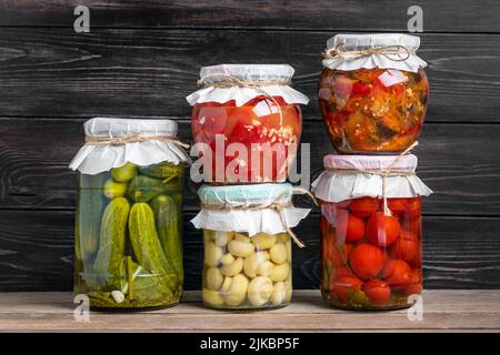 Hausgemachte eingelegte Kirschtomaten, Gurken, Champignons, Knoblauch, Auberginen, Rote Paprika in Gläsern auf Holzregal hausgemachte Konserven und fermentierte Lebensmittel Konzept Saisonprodukt. Stockfoto