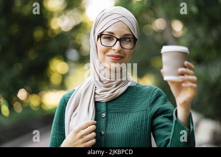 Porträt einer jungen muslimischen Frau, die einen Kopftuch mit Hijab und Kaffee trägt, um in den Stadtpark zu gehen. Nahaufnahme der fröhlichen Frau, bedeckt mit Kopftuch und einem heißen Getränk, das im Freien lächelt. Stockfoto