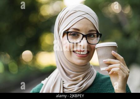 Porträt einer jungen muslimischen Frau, die einen Kopftuch mit Hijab und Kaffee trägt, um in den Stadtpark zu gehen. Nahaufnahme der fröhlichen Frau, bedeckt mit Kopftuch und einem heißen Getränk, das im Freien lächelt. Stockfoto