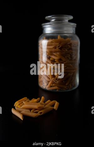 Vollkorn-Penne-Pasta vor dem Glasglas auf schwarzem Hintergrund Stockfoto