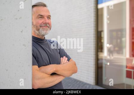 Ein entspannter Mann mittleren Alters, der mit einem amüsierten Lächeln steht und sich mit Copyspace an eine Wand lehnt Stockfoto