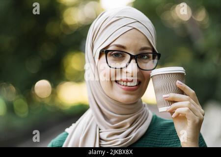Porträt einer jungen muslimischen Frau, die einen Kopftuch mit Hijab und Kaffee trägt, um in den Stadtpark zu gehen. Nahaufnahme der fröhlichen Frau, bedeckt mit Kopftuch und einem heißen Getränk, das im Freien lächelt. Stockfoto