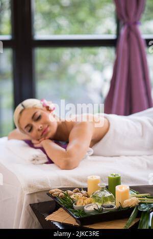 Hübsche blonde Frau, die nach einer angenehmen Spa-Behandlung auf dem Massageliege schlief, die Inneneinrichtung des Salons im Hintergrund Stockfoto