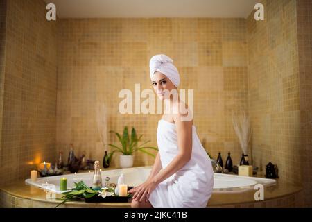 Attraktive junge Frau, die ein Handtuch trägt und die Kamera anschaut, während sie am Rand der Badewanne sitzt, die Inneneinrichtung des modernen Badezimmers im Hintergrund Stockfoto