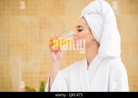 Profilansicht einer hübschen jungen Frau, die Bademantel und Handtuch trägt und frisch gepressten Saft trinkt, während sie das Wochenende im Spa-Salon genießt, verschwommener Hintergrund Stockfoto