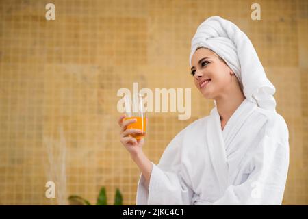 Porträtaufnahme einer lächelnden jungen Frau, die einen Bademantel und ein Handtuch trägt und ein Glas frisch gepressten Saft hält und wegschaut, im Hintergrund die Inneneinrichtung des Spa-Salons Stockfoto