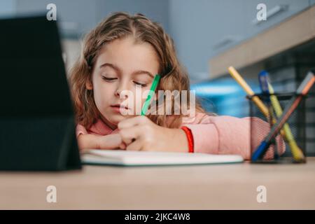 Schulmädchen beobachten Online-Kurs auf Tablet in der Küche, Notizen machen, Hausaufgaben machen. Virtuelle Unterrichtsstunde auf Video während der Selbstisolation zu Hause Stockfoto