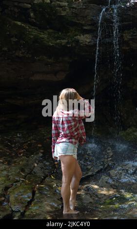 Schöne junge Frau in abgeschnittenen Jeans und rotem Flanellhemd, die unter einem Wasserfall über die Felsen läuft Stockfoto