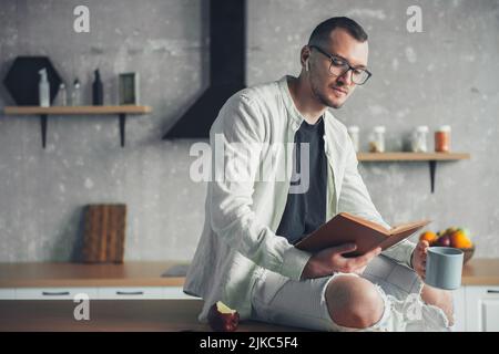 Seriöser junger Geschäftsmann trinkt Morgenkaffee in der Küche und liest ein Buch. Unternehmensporträt. Lesen Sie ein Papierbuch und trinken Sie morgens Kaffee Stockfoto
