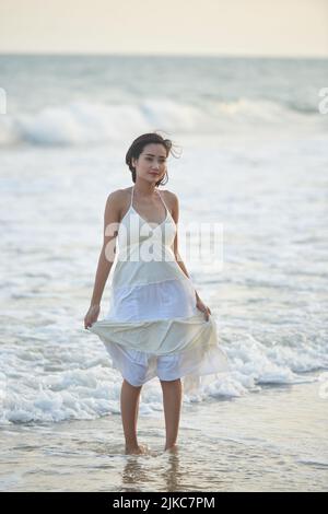 In voller Länge Porträt von attraktiven asiatischen Frau trägt Sommerkleid weg pensive suchen, während knöcheltief im Wasser des welligen Meeres stehen Stockfoto