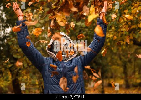 Unschärfen Sie Halloween-Menschen. Person in grimmiger Reaper Maske, die die Hand hebt und Blätter wirft. Viele fliegende orange, gelbe, grüne trockene Blätter. Nicht fokussiert. Stockfoto