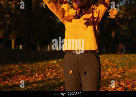 Unschärfe hallo Herbst. Glückliche blonde elegante 40-jährige Frau in gelbem Pullover, Schal draußen genießen Herbst und werfen Blätter. Viele fliegen orange Stockfoto