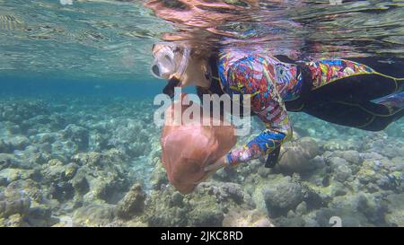 Frau in Tauchausrüstung schwimmt und sammelt Plastikmüll unter Wasser auf dem Boden des Korallenriffs. Schnorchler reinigt Ozean von Plastikverschmutzung. P Stockfoto
