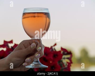 Eine Nahaufnahme der Hand einer Frau, die ein Weinglas mit verschwommenen roten Blumen im Hintergrund hält Stockfoto