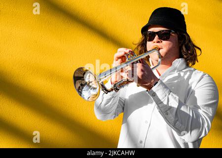 Junger kaukasischer langhaariger Mann in Hemd und Sonnenbrille, der draußen Trompete spielt Stockfoto