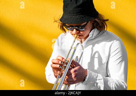 Junger kaukasischer langhaariger Mann in Hemd, Hut und Sonnenbrille, der Trompete spielt Stockfoto