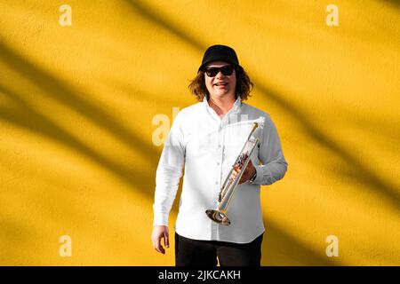 Der junge kaukasische langhaarige blonde Mann in Hemd und Hut spielt im Freien Trompete Stockfoto