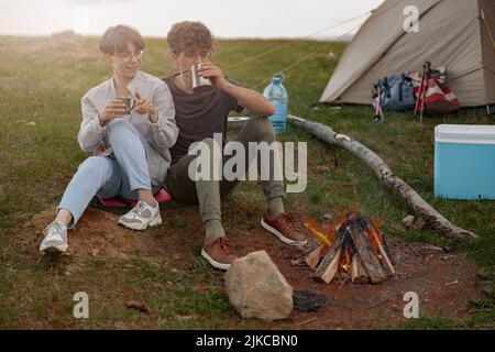 Junge Touristen sitzen am Feuer im Lager und trinken heiße Getränke. Stockfoto