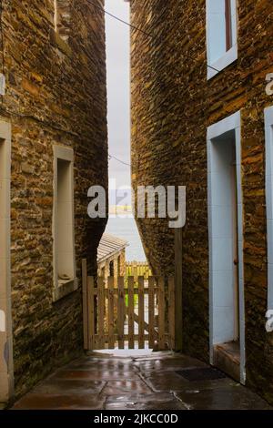 Eine vertikale Aufnahme einer schmalen Straße mit einem Holzzaun und einem Meer im Hintergrund in Orkney, Schottland Stockfoto
