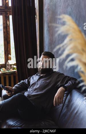 Ein Mann mit Bart und Smartphone sitzt auf einem Ledersofa. Stockfoto