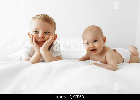 Zwei glückliche Kinder liegen auf einem weißen Bett in einem Zimmer. Netter älterer Bruder mit einem neugeborenen Baby. Glücklicher Bruder und Schwester spielen zusammen im Bett, glückliche Familie Stockfoto