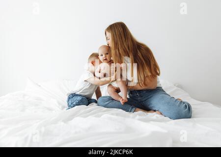 Glückliche Mutter mit zwei kleinen Kindern sitzt auf einem weißen Bett im Schlafzimmer, glückliche Mutter umarmt und küsst ihre Kinder, schaut einander an, Familie genießt es, zu pflegen Stockfoto