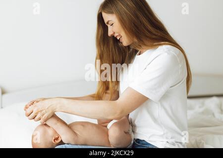 Happy Baby in Windel bekommt von der Mutter zu Hause auf weißem Bett eine Bauchmassage. Babymassage Mutter massiert ihren Bauch, das Kind lacht. Behandlung der Kolonne Stockfoto