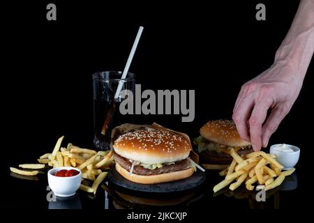 Männliche Hand Dipping pommes in Pilzsauce. Frisch leckeren Hamburger, pommes frites, Cola auf schwarzem Glas mit Reflexion, selektiver Fokus, Seitenansicht Stockfoto